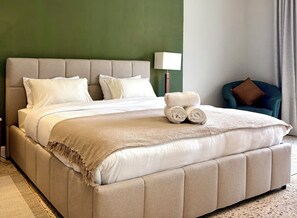 Una cama con cabecera color beige, ropa de cama blanca y toallas enrolladas.