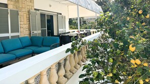 A covered outdoor seating area with a white balustrade, a stone wall, and a view of a garden with trees and plants.