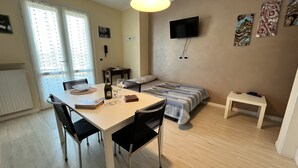 Ein gemütliches Hotelzimmer mit Esstisch, Stühlen, einem Bett mit gestreifter Decke, einem kleinen Nachttisch, einem an der Wand montierten Fernseher und Bildern an den Wänden.
