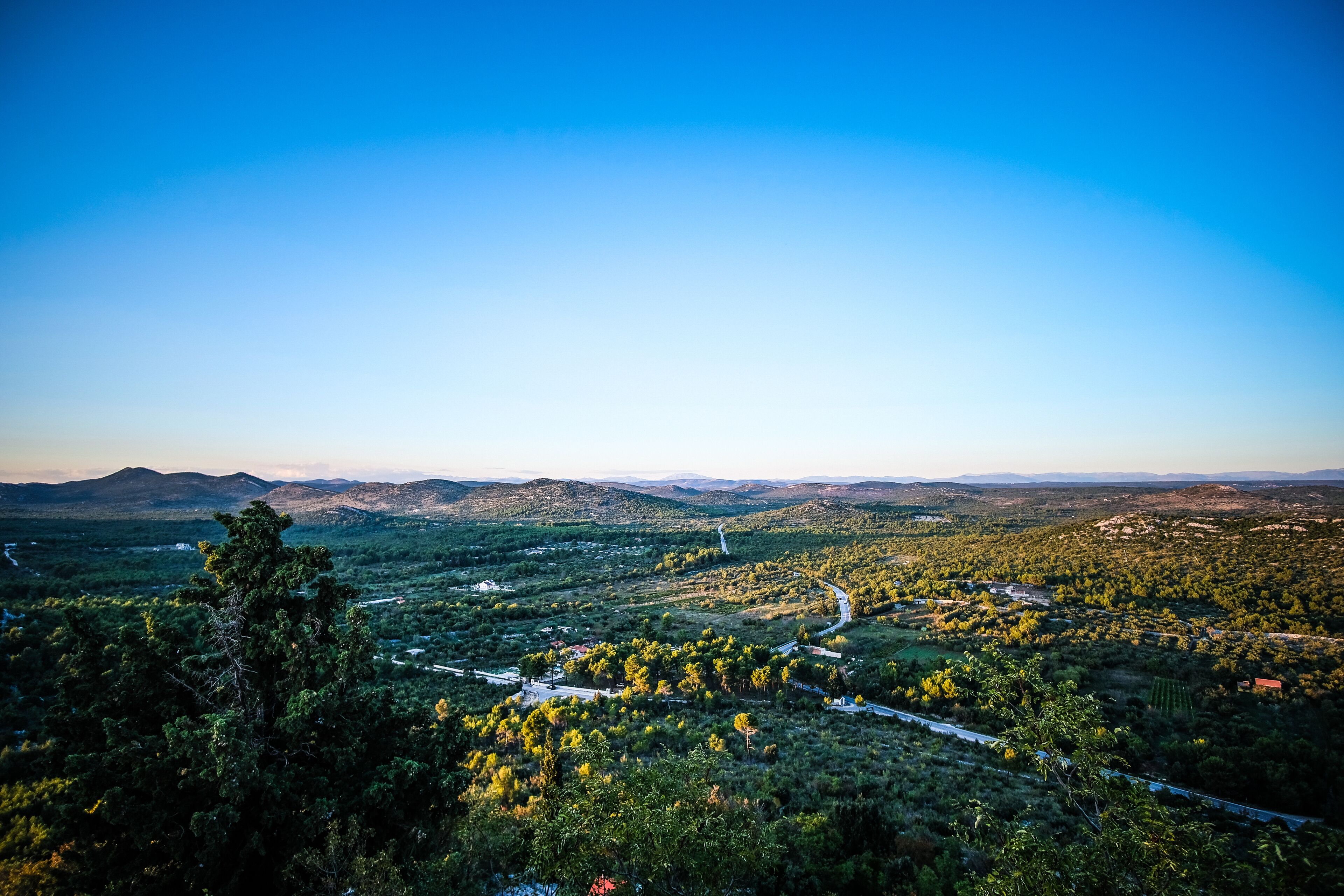 A panoramic view of a mountainous landscape with a winding road, dense vegetation, and distant hills.