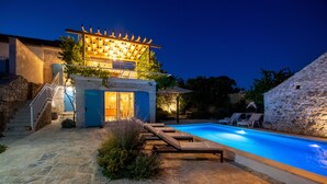 A stone building with a pool, lounge chairs, and a pergola with lights.