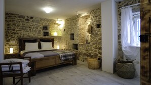 A stone-walled bedroom with a wooden bed, a chair, a bedside table with a lamp, and a window with a curtain.