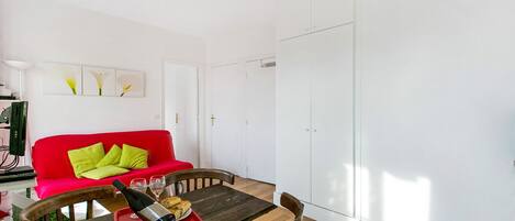 Un coin repas avec une table en bois, des chaises rouges et vertes, un plateau rouge avec une bouteille et des verres, et un canapé rouge avec des coussins verts.