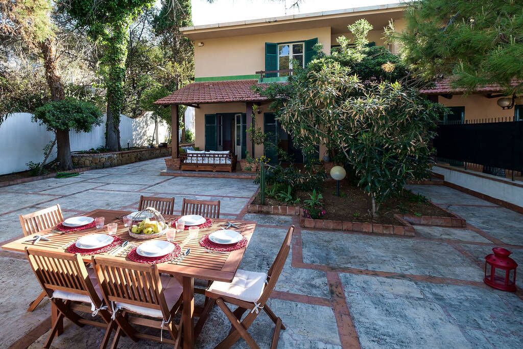 A patio with a wooden table set for dining, surrounded by chairs, and a house in the background.