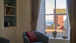 Ein Zimmer mit einem großen Fenster, einem blauen Sessel, einem Bücherregal und Blick aufs Meer.