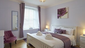 Ein Schlafzimmer mit einem weißen Bett, violetter Bettwäsche, einem Spiegel, einem Fenster mit Vorhängen und einem Nachttisch mit einer Lampe.