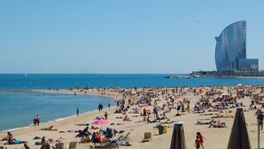 A crowded beach with people sunbathing and swimming. A sailboat is visible in the distance. - Discover Comfort & Culture in the Heart of Barcelona! (Barcelona)