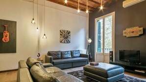 A modern living room with a sectional sofa, a black ottoman, a TV, and a guitar decoration on the wall. - Discover Comfort & Culture in the Heart of Barcelona! (Barcelona)