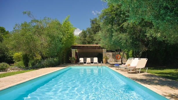 Una piscina rectangular con agua azul cristalina, rodeada por una terraza de piedra. Hay sillas reclinables blancas y un área de estar cubierta con un muro de piedra y un techo de azulejos.