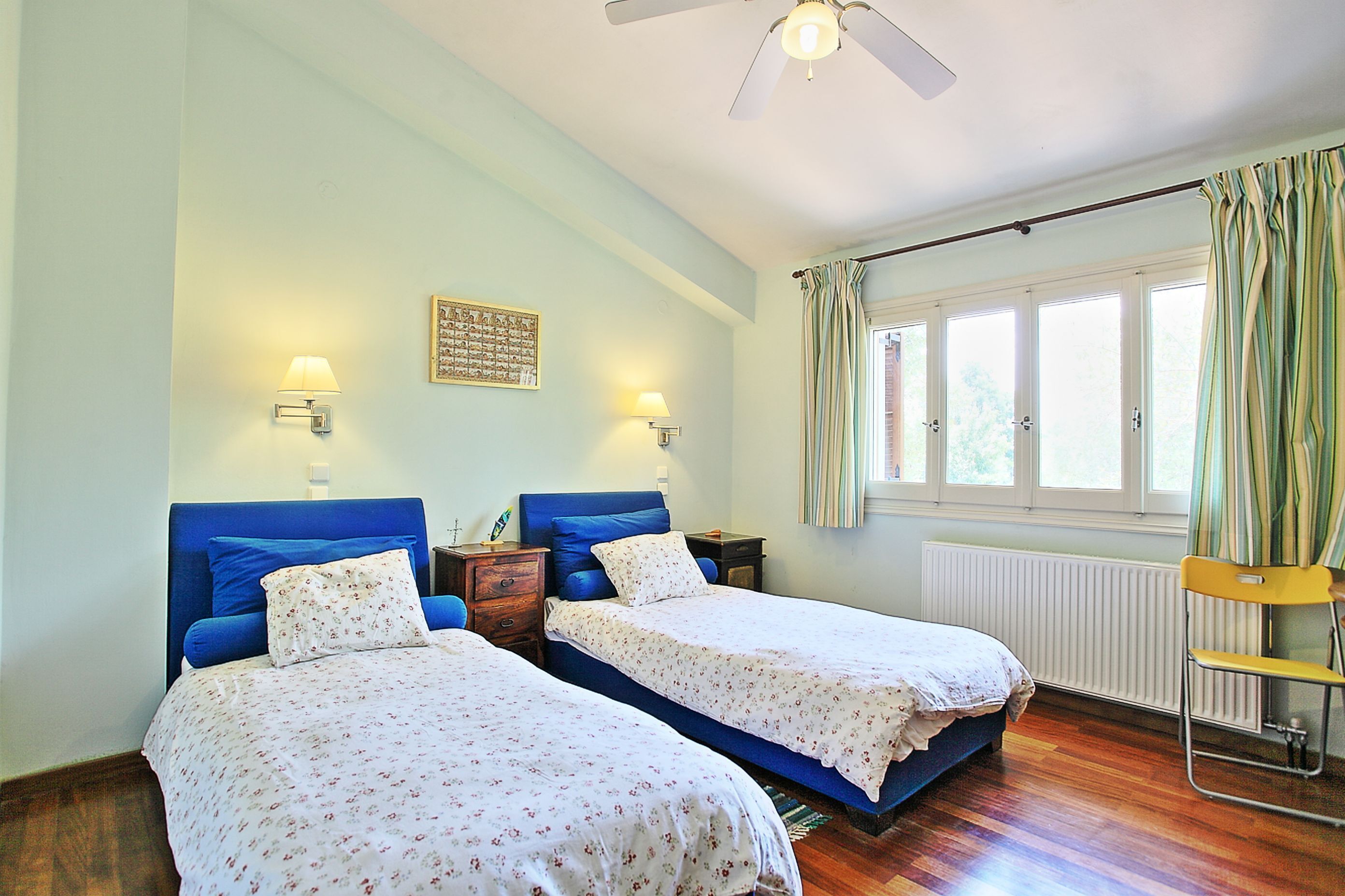 Een slaapkamer met twee bedden, een plafondventilator, een raam met gordijnen en een radiator.