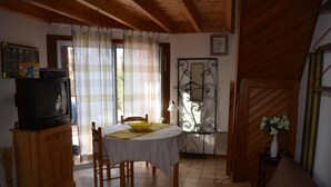 Un coin repas comprenant une table, des chaises, un téléviseur posé sur un meuble et une fenêtre avec des rideaux.