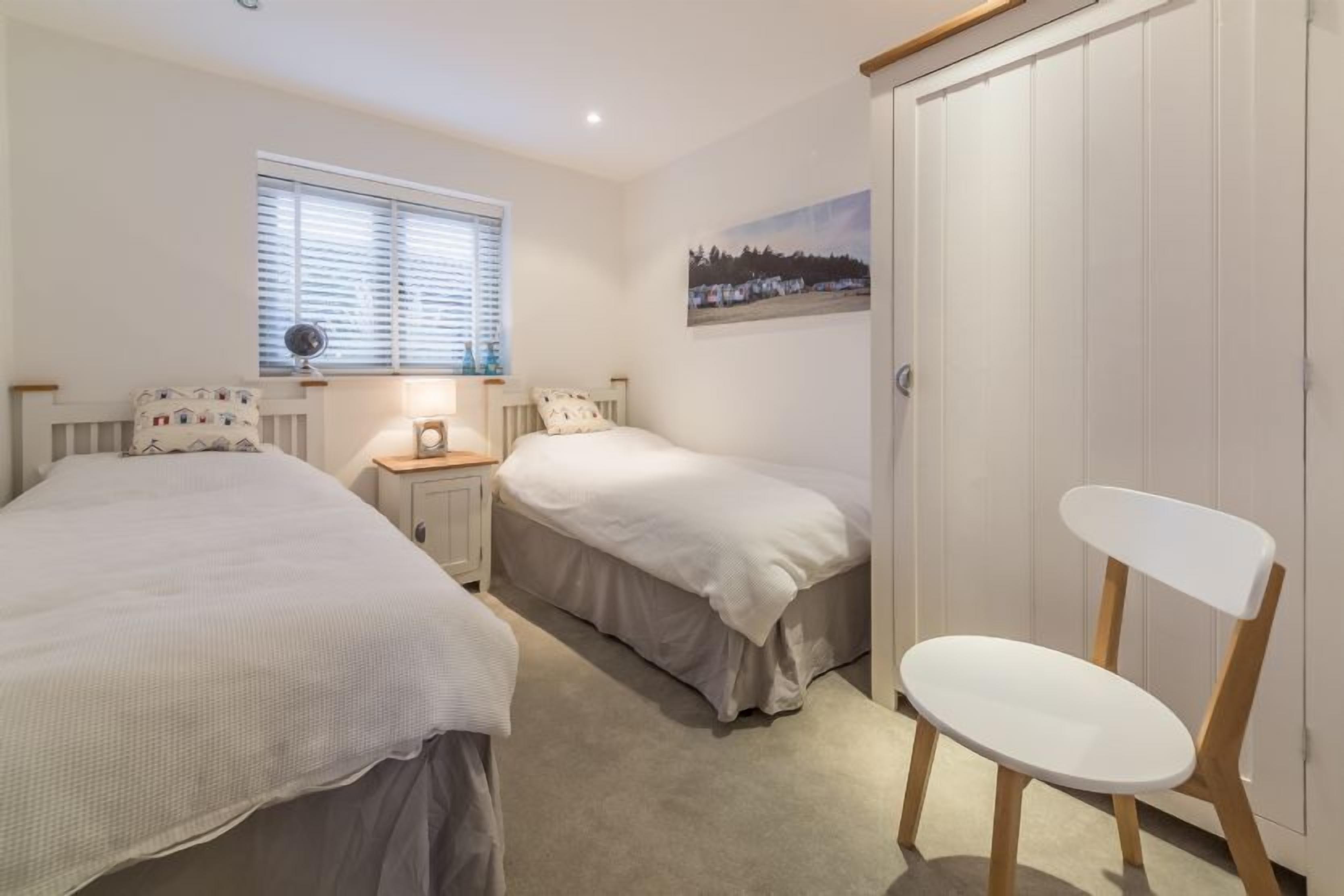 A bedroom with two beds, a bedside table with a lamp, a chair, a window with blinds, and a door.