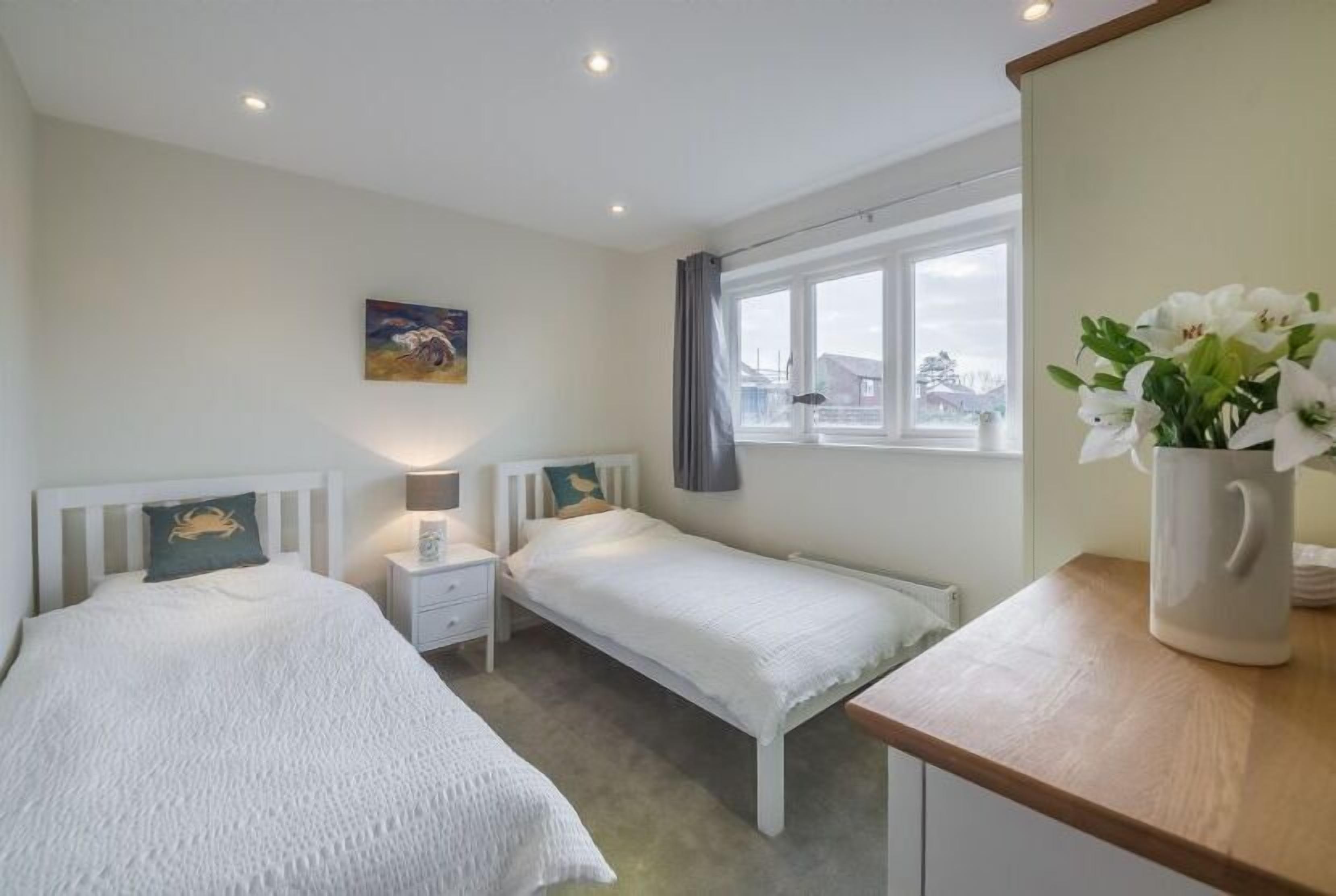 A bedroom with two single beds, a nightstand with a lamp, a window with curtains, and a vase with flowers on a wooden table.