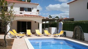A swimming pool with sun loungers, a covered patio, and a white building with a tiled roof.
