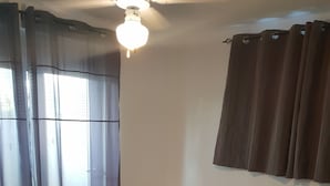Une chambre d’hôtel avec deux lits, chacun recouvert d’une couverture sombre et agrémenté d’une serviette blanche roulée ornée de fleurs orange. On y trouve un ventilateur de plafond, une fenêtre avec des rideaux et un luminaire fixé au mur.