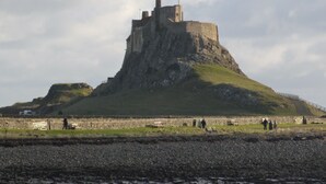 Eine Burg auf einem Hügel mit einer grünen Wiese im Vordergrund und einer felsigen Küste.