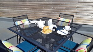 Ein quadratischer Tisch im Freien mit vier Stühlen, jeder mit bunten, gestreiften Kissen. Auf dem Tisch stehen Teller, Tassen, ein Teekessel und Croissants.