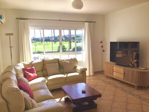 Un salon avec un canapé beige, une table basse en bois, un téléviseur posé sur un meuble en bois, et une vue sur un paysage verdoyant grâce à de grandes fenêtres.