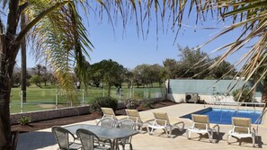 Un espace piscine doté de chaises longues et d’une table, entouré de palmiers et offrant une vue sur un terrain de golf.