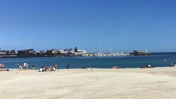 Una playa con gente tomando el sol y nadando, un muelle con barcos, y edificios al fondo.