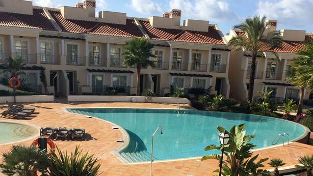 A swimming pool with a tiled deck, surrounded by palm trees and a building with a tiled roof.