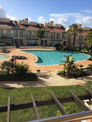 A swimming pool with a tiled deck, surrounded by palm trees and a building with a tiled roof. - Palmyra, Vila Sol, Vilamoura (Quarteira)