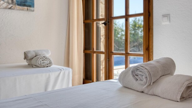A room with a bed, a window with wooden frames, a painting of birds, and a table with towels.