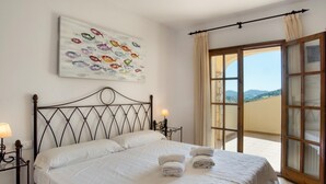 A neatly made bed with white linens and towels, a painting of fish on the wall, and a balcony with a view.