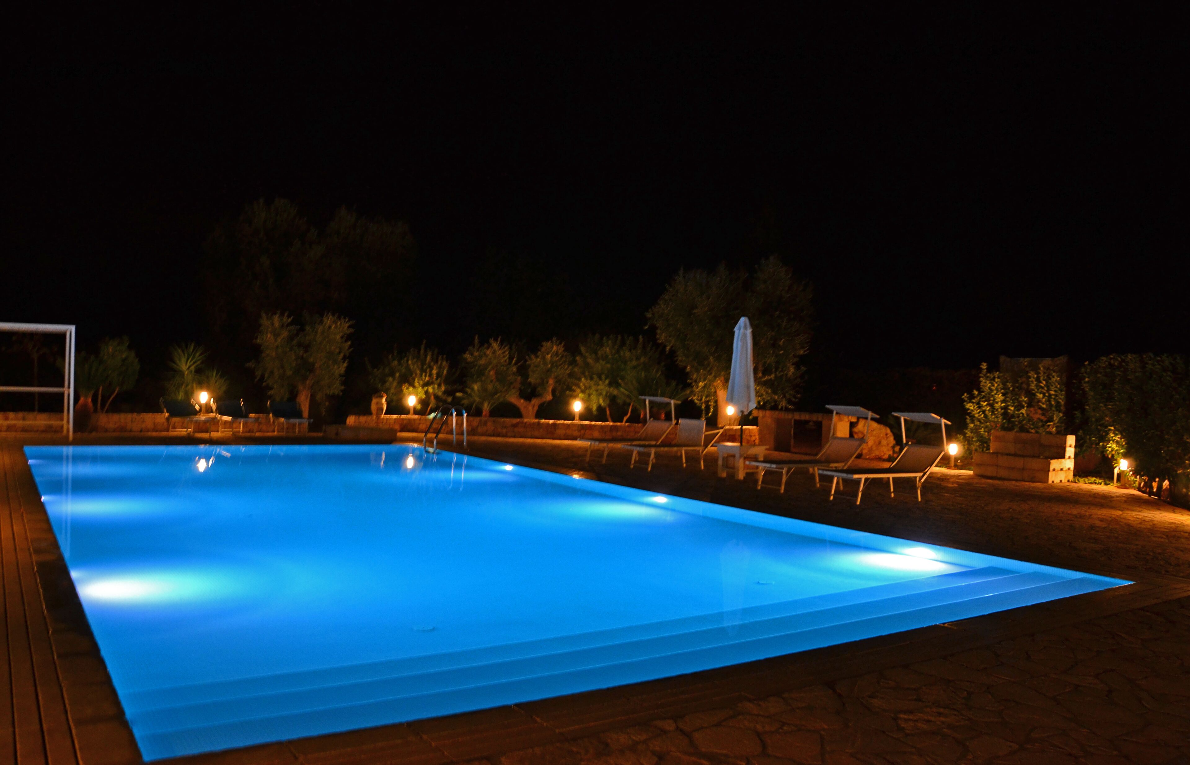 A swimming pool at night with illuminated edges, surrounded by lounge chairs and a pergola.
