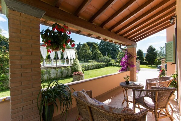 Une terrasse couverte, meublée de mobilier en osier, avec vue sur un jardin et un mur en briques.