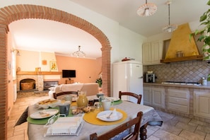 Un coin repas chaleureux agrémenté d’une arche en briques, d’une cheminée et d’une cuisine avec des meubles blancs et une hotte jaune.