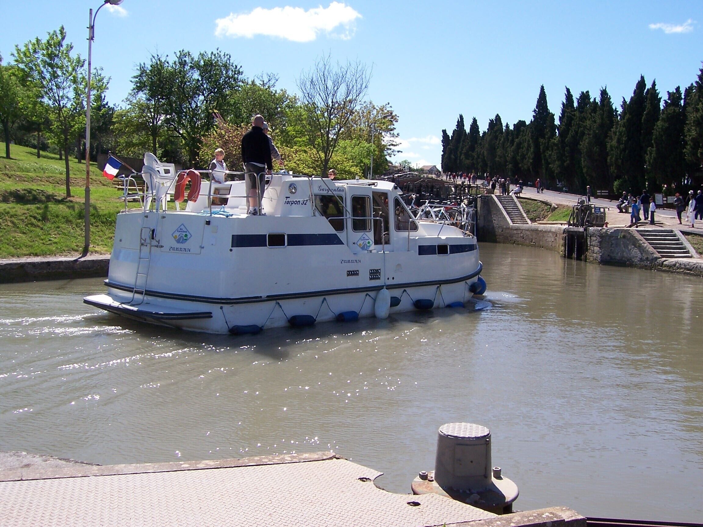 A white boat with a cabin and a small deck, docked at a pier with a crowd in the background.