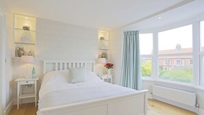 A bedroom with a white bed, bedside tables, a window with curtains, and shelves with decorative items.