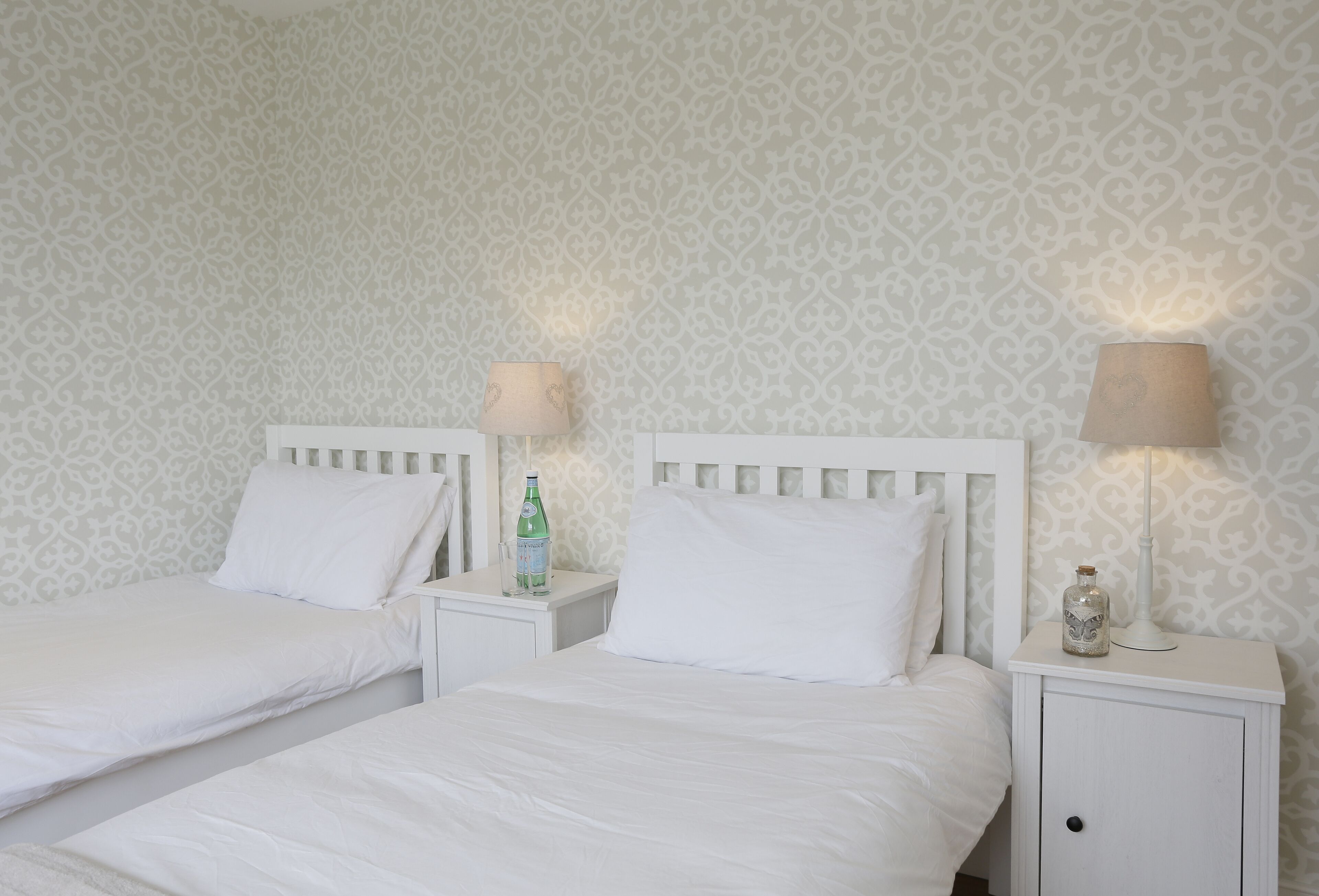 Une chambre d’hôtel avec deux lits simples, des têtes de lit blanches, des tables de chevet avec des lampes et un papier peint à motifs.