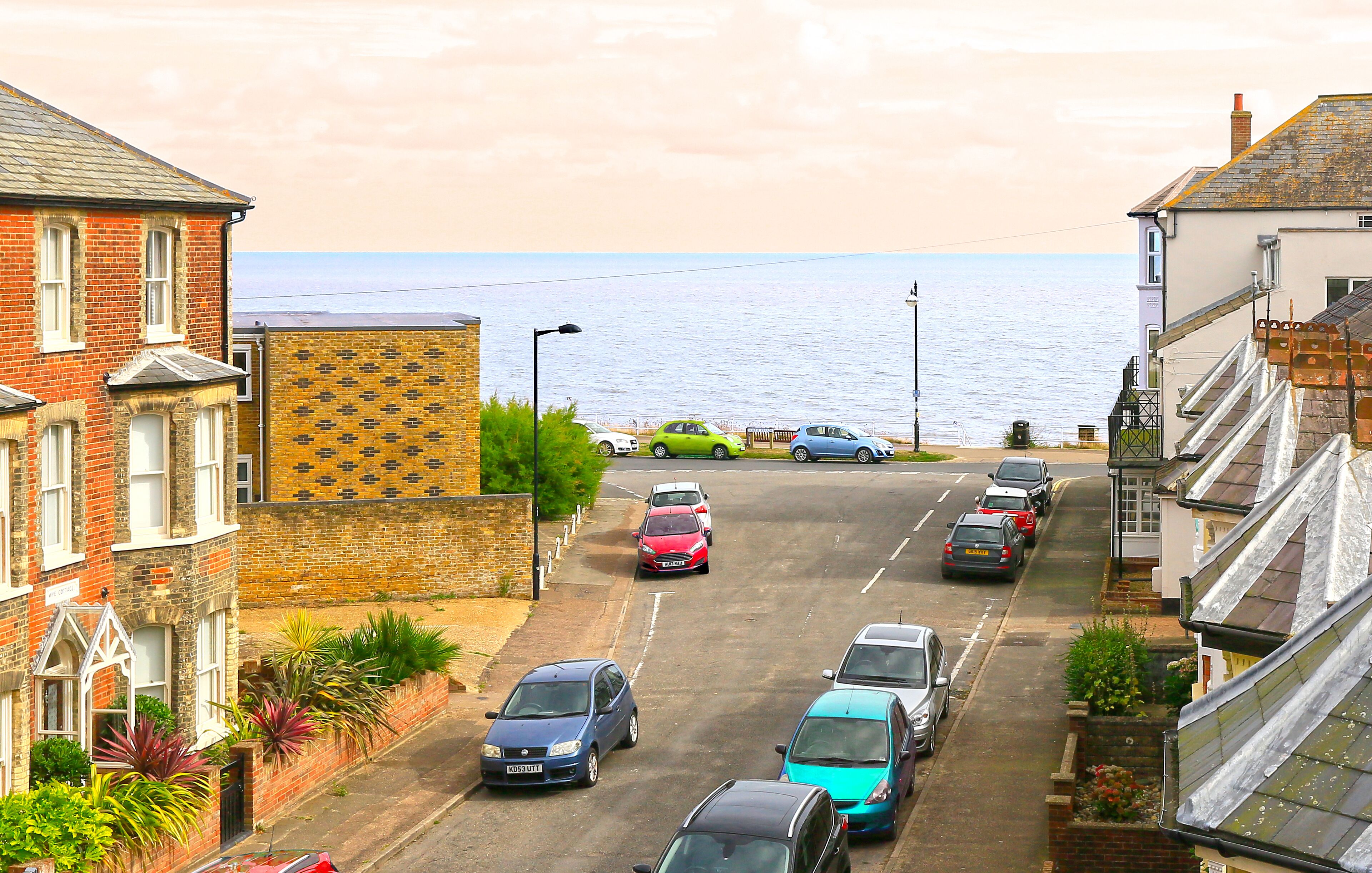 Una calle costera con coches aparcados, un edificio de ladrillo con la inscripción 'KYE COTTAGE' y vistas al mar.