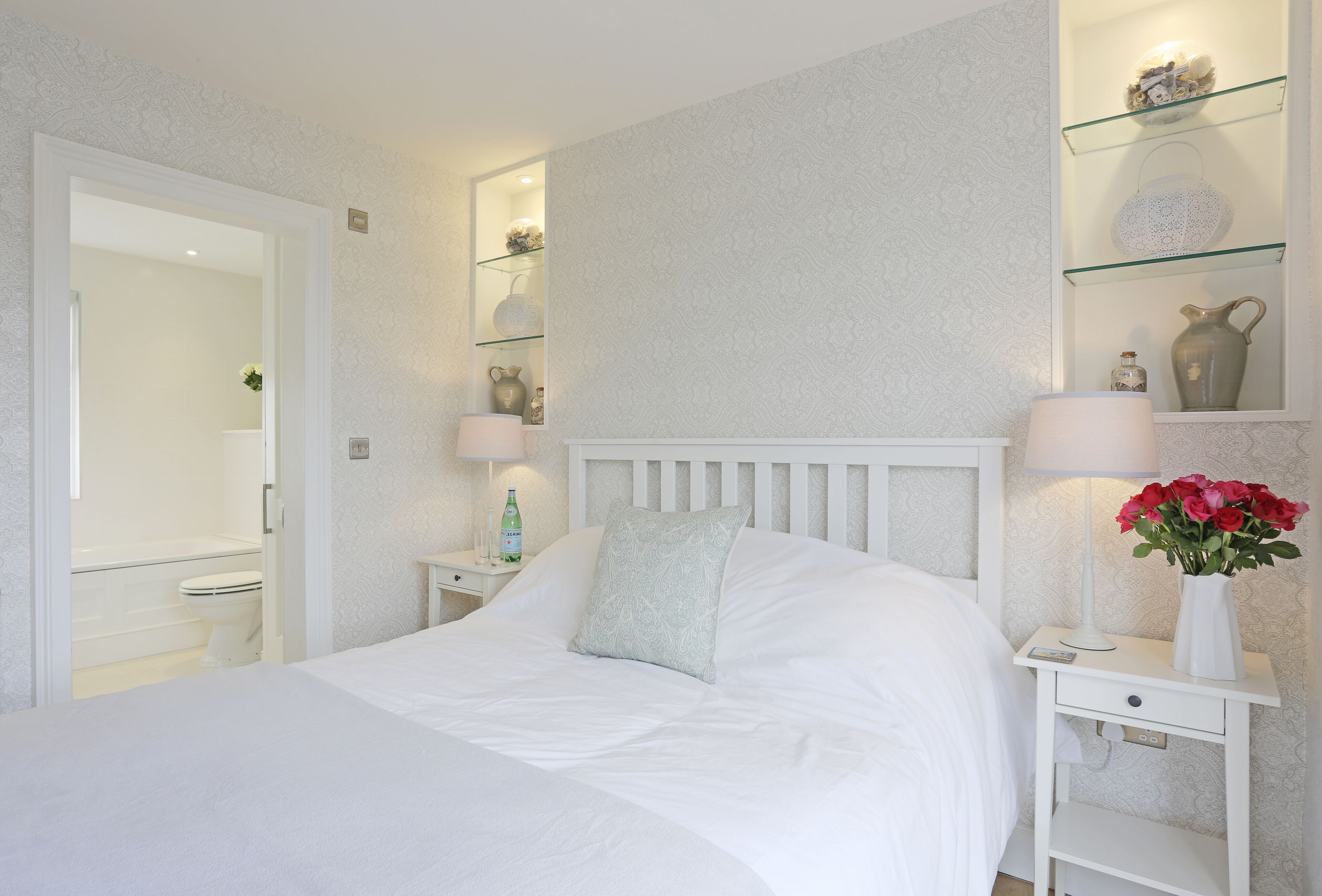 A neatly arranged bedroom with a bed, bedside tables, a nightstand, and a glass shelf with decorative items.