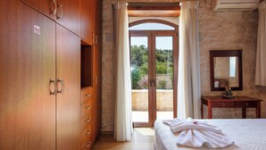 A bedroom with a wooden wardrobe, a bed with a white duvet, a wooden nightstand, and a view of greenery through a glass door. - Stone Made villa in picturesque village! Private pool & full privacy! (Rethymno)
