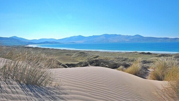 Paisatge costaner amb dunes de sorra i una vista clara de l'oceà i muntanyes allunyades.