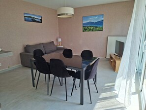 Een moderne woonkamer met een eettafel, zwarte stoelen, een grijze bank, een televisie en twee landschapsschilderijen aan de muur.