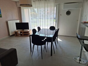 Een moderne woonkamer met een glazen eettafel, zwarte stoelen, een televisie op een houten dressoir en een groot raam met dunne gordijnen.