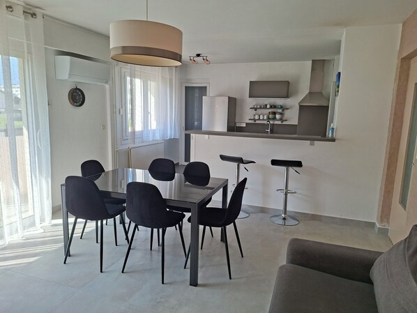 Een moderne keuken met een inbouwoven en magnetron, een eethoek met een glazen tafel en zwarte stoelen, en een zithoek met een bank.