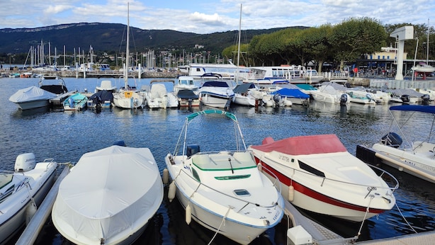 Un port de plaisance où sont amarrés différents types de bateaux, un ciel dégagé et un paysage montagneux en arrière-plan.