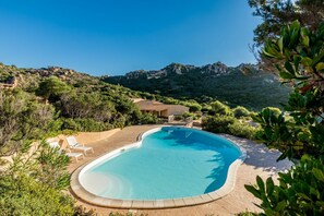En swimmingpool med buet kant, omgivet af liggestole og grønt, med et bjergrigt landskab i baggrunden.
