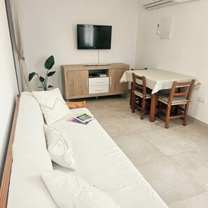 Una sala de estar con sofá, una mesa de comedor con sillas, un televisor montado en la pared y un aparador.