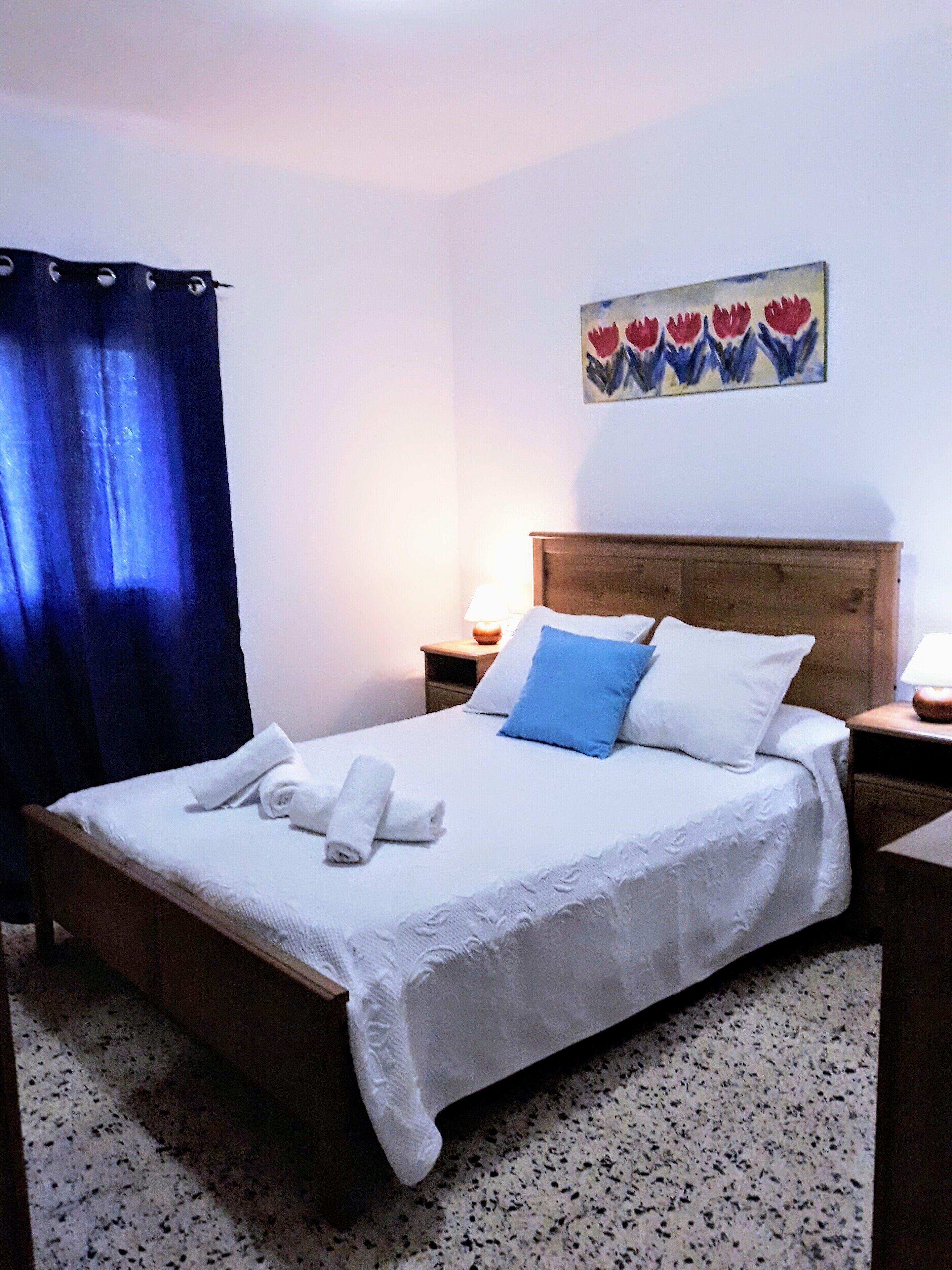 A neatly made bed with white linens and rolled towels, a wooden headboard, and a painting of flowers on the wall.