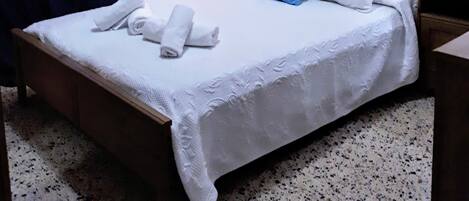 A neatly made bed with white linens and rolled towels, a wooden headboard, and a painting of flowers on the wall.