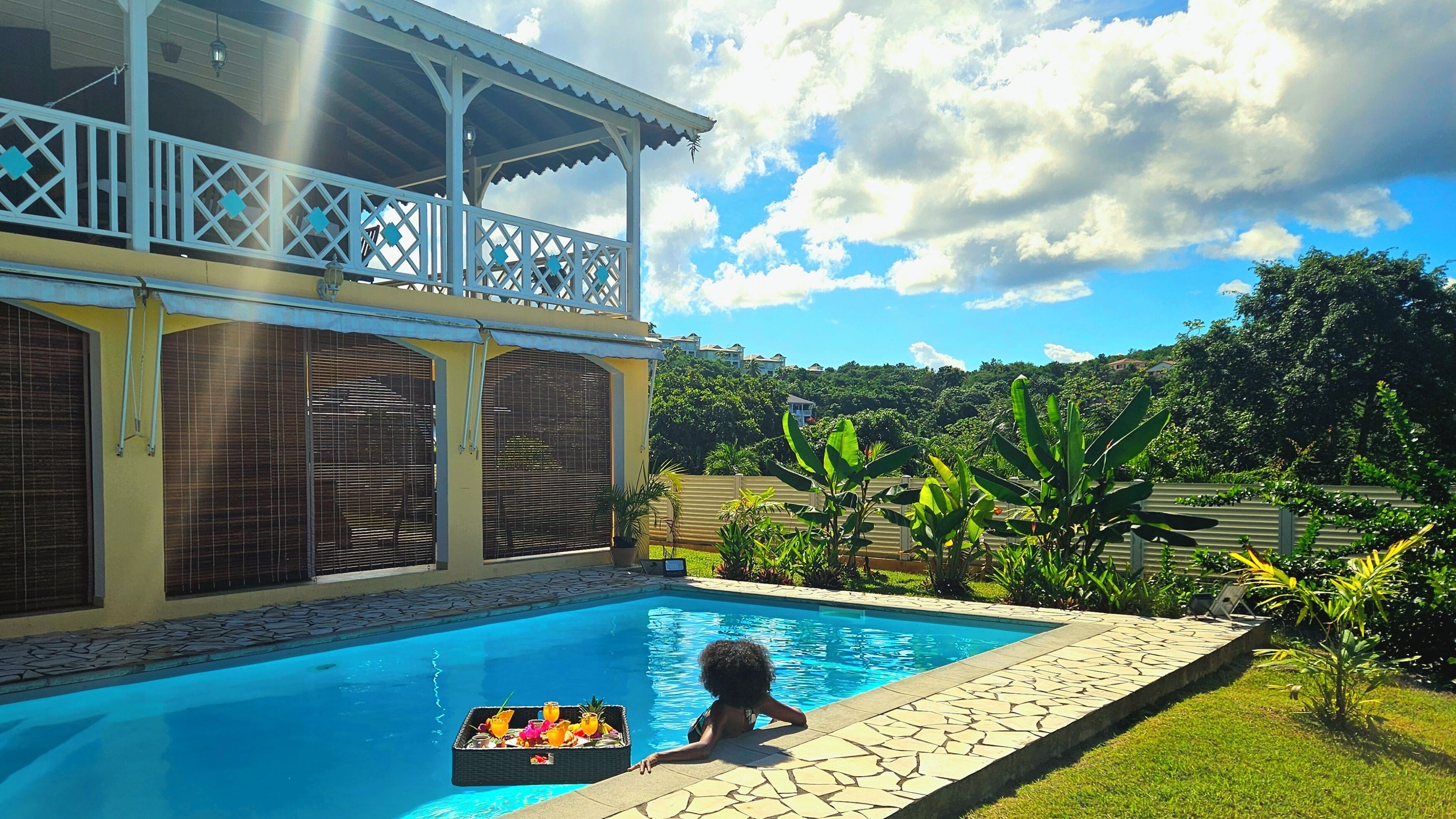 A swimming pool with a child playing, surrounded by lush greenery and a covered patio area.