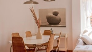 Zona pranzo con tavolo rotondo, sedie in vimini, un vaso con piante essiccate e un quadro appeso.