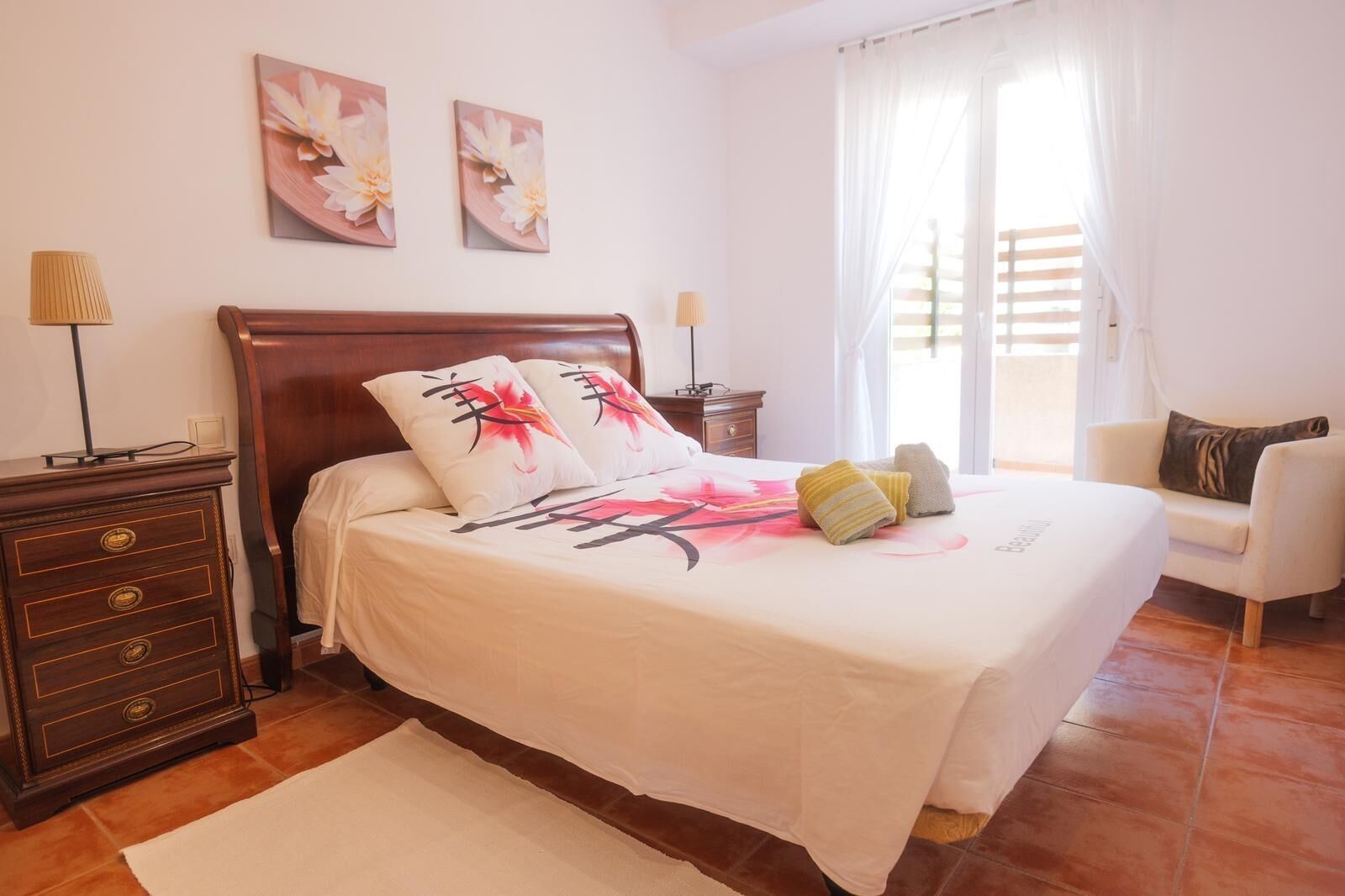 Une chambre à coucher bien rangée, avec un lit en bois, deux lampes fixées au mur, une table de chevet et une chaise.