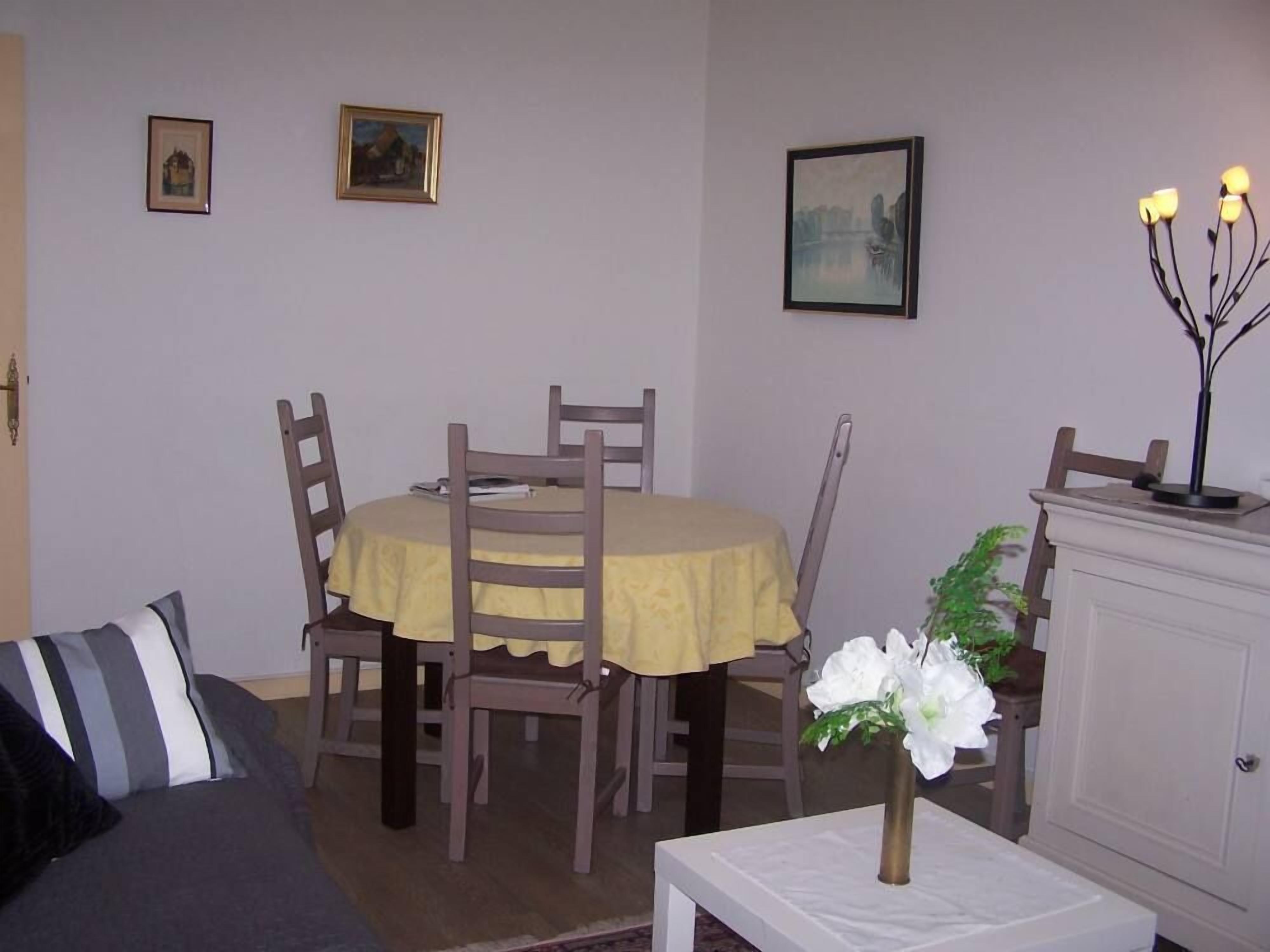 Uma área de refeições com uma mesa redonda coberta por uma toalha amarela, rodeada por cadeiras de madeira. Há três quadros na parede, um armário branco à direita e uma pequena mesa com um vaso de flores em primeiro plano.